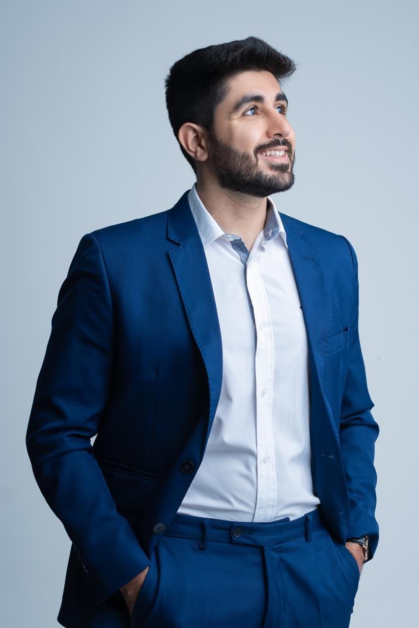 Dr. Ali Hamie sorrindo, de terno azul, olhando para o lado em retrato de estúdio.
