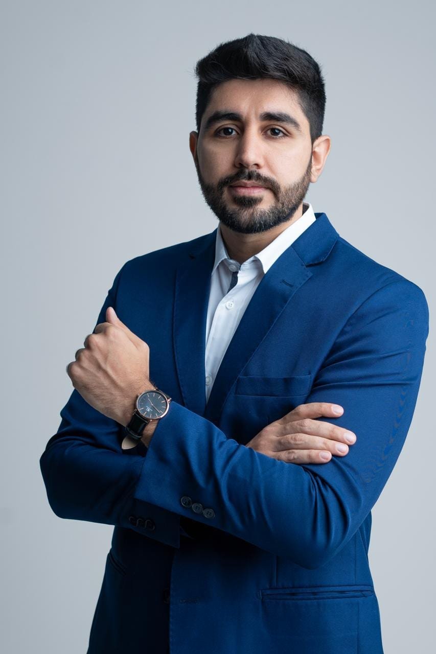 Dr. Ali Moussa Hamie Neto, de terno azul e braços cruzados, em retrato de estúdio.